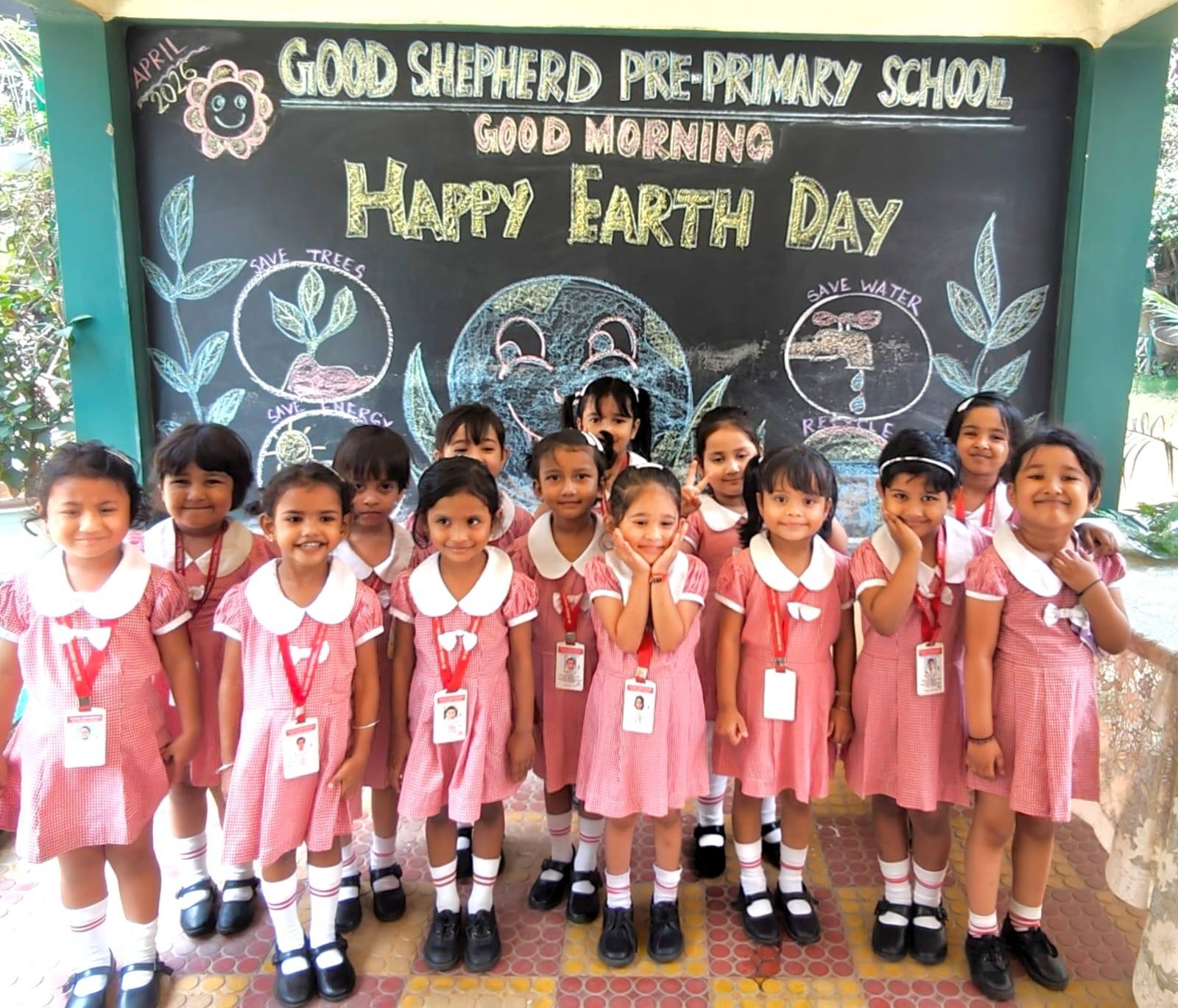 A happy group of girls in pink checked uniforms in front of a chalkboard reading “Good Shepherd Pre-Primary School – Good Morning – Happy Earth Day” with save trees and save water drawings, Guwahati