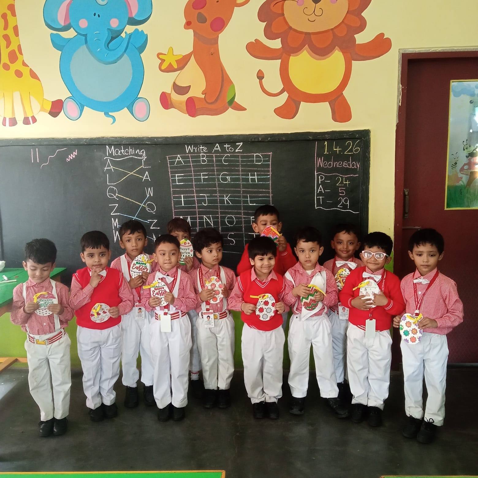 Boys in red and white uniforms hold up their handmade Easter egg cards in a classroom with an A to Z chalkboard and painted animal murals