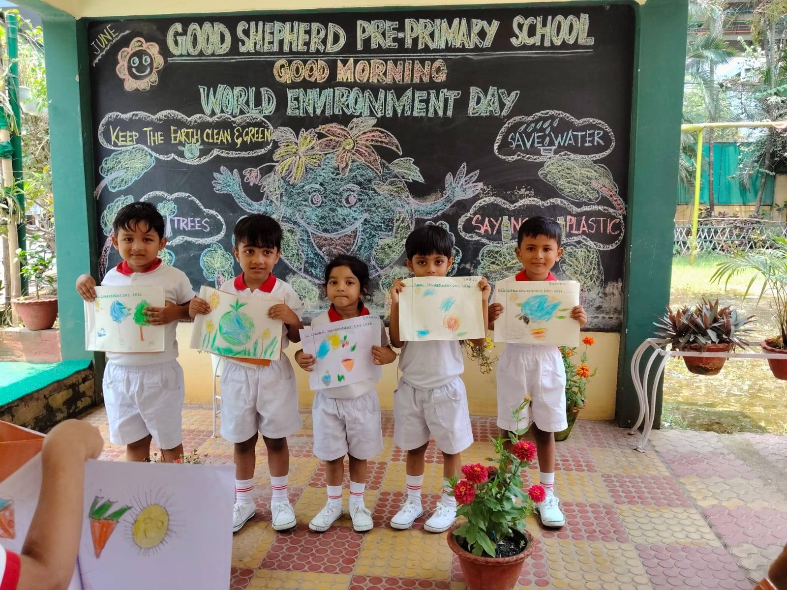 Five children in white uniforms show their crayon drawings in front of a “World Environment Day” chalkboard on the school courtyard in Chandmari, Guwahati