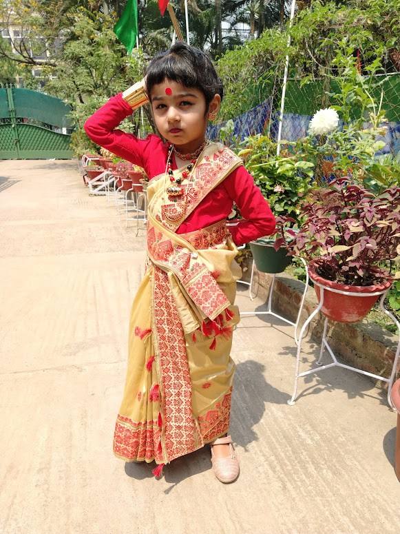 A little girl in a traditional Assamese mekhela chador poses on a paved path lined with potted plants