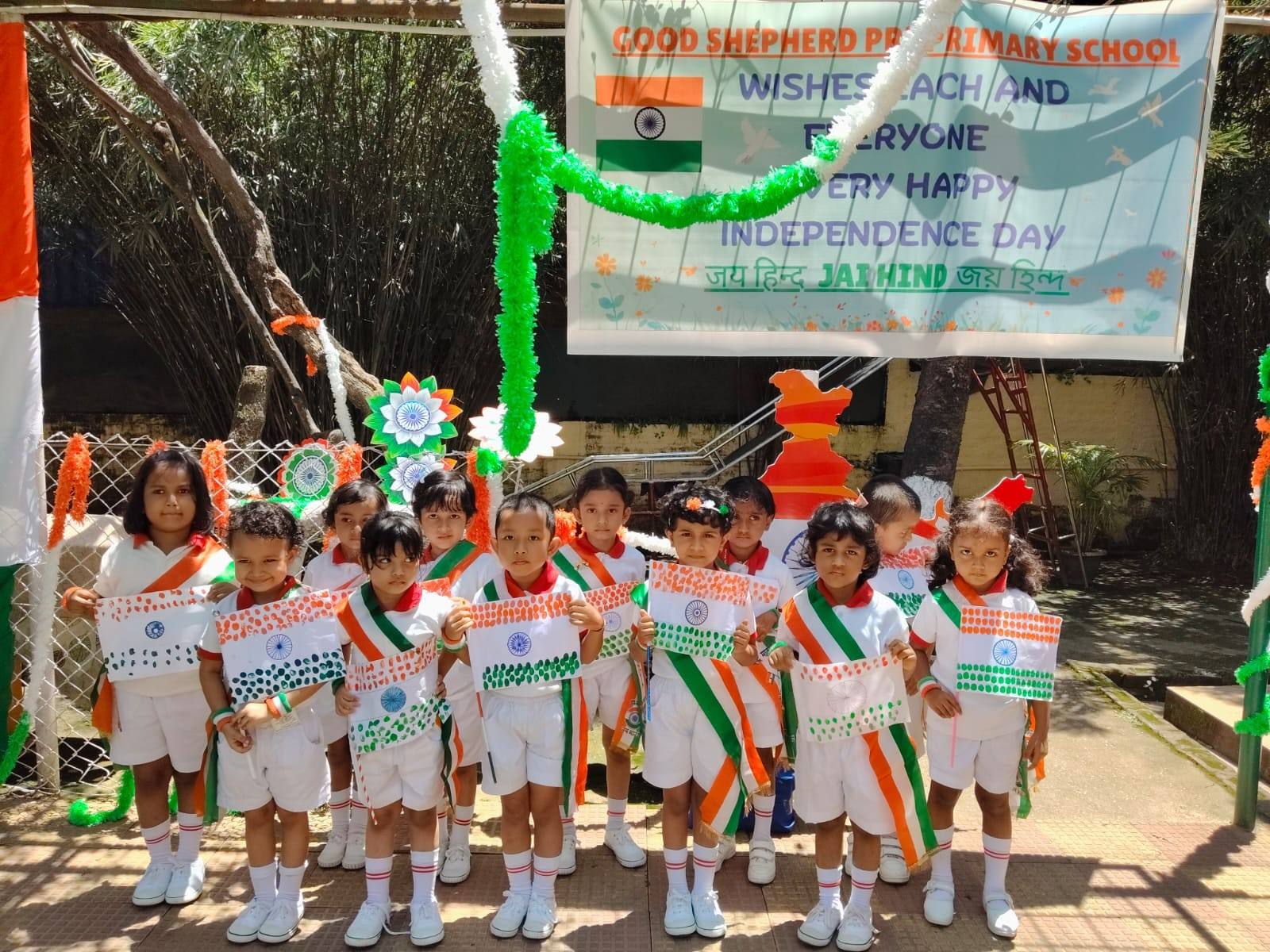 Children wearing tricolour sashes hold hand-painted Indian flags under an Independence Day banner and tricolour decorations at Good Shepherd Pre Primary School, Guwahati