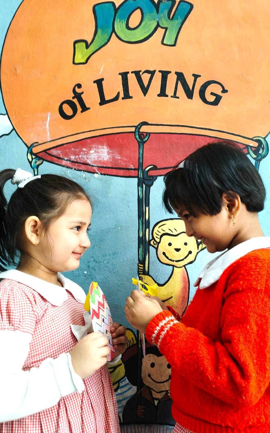Two little girls smile at each other while holding their paper crafts in front of a painted “The Joy of Living” hot-air balloon mural at school